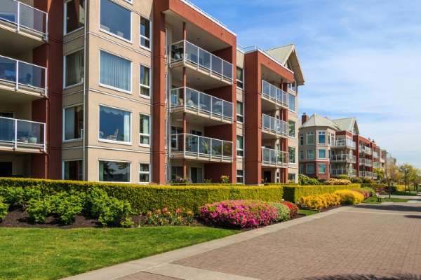 Apartment complex exterior with balconies, landscaped hedges, and a paved walkway under a blue sky